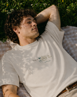 A man lies on a checkered blanket in a grassy outdoor setting, wearing a white Christian t-shirt and beige pants. A basket of white flowers and a bottle are visible next to him.